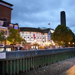 Kvällsutsikt över en byggnad vid floden med arkitektur i Tudor-stil, träd och människor som går på strandpromenaden.
