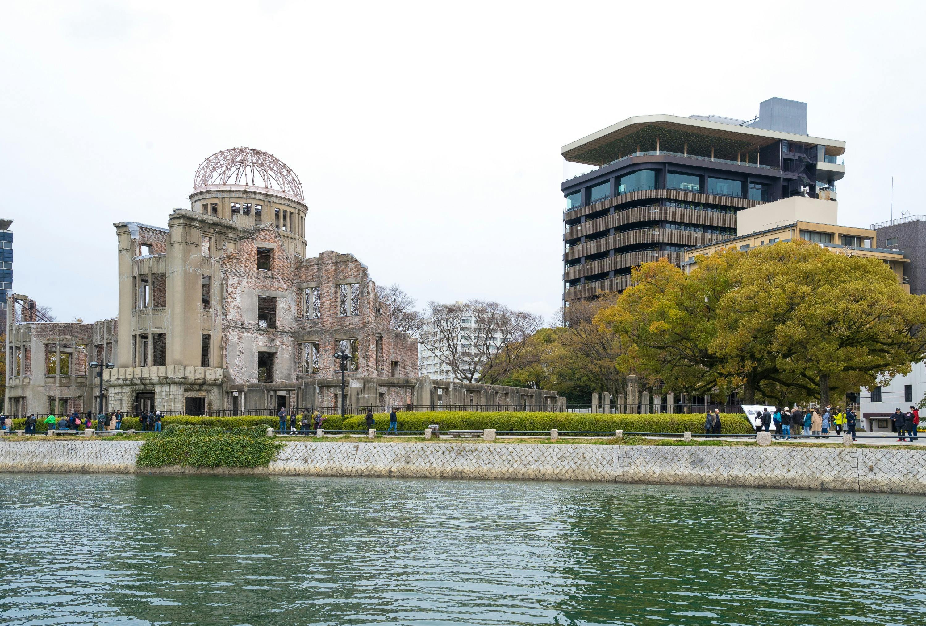 A riverside view featuring the Hiroshima Peace Memorial and modern buildings, with trees and people walking along the path.
