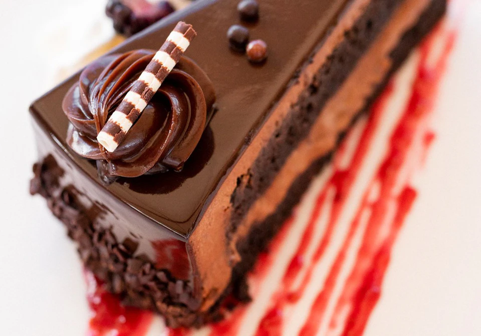 A slice of chocolate cake with glossy icing, chocolate curls, and striped chocolate stick on top, set on a plate with red sauce drizzle.