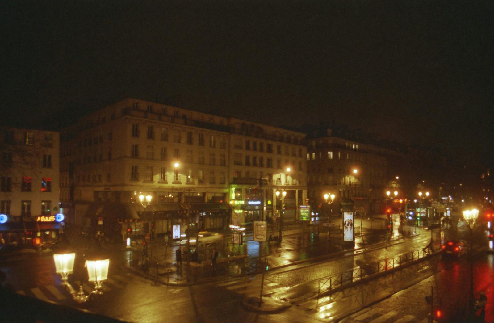 Nocą ulica miejska z mokrym chodnikiem, budynkami, latarniami ulicznymi i podświetlanymi znakami sklepowymi. Niewiele osób i samochodów jest widocznych.