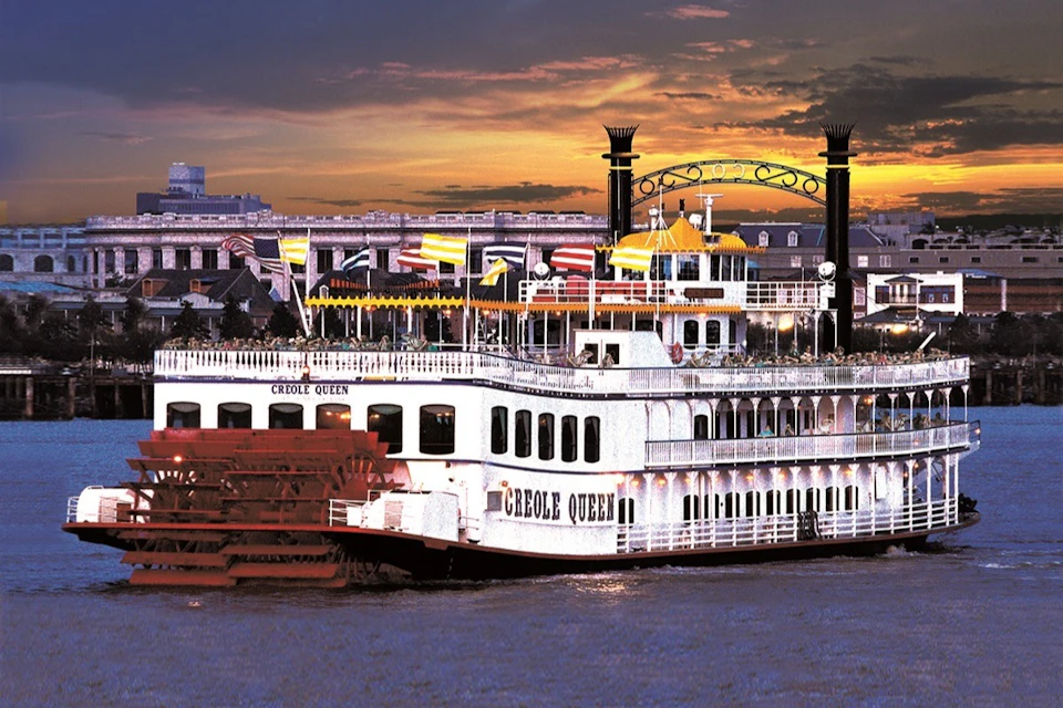 A riverboat named "Creole Queen" with multiple decks and colorful flags sails at sunset, with a city backdrop.