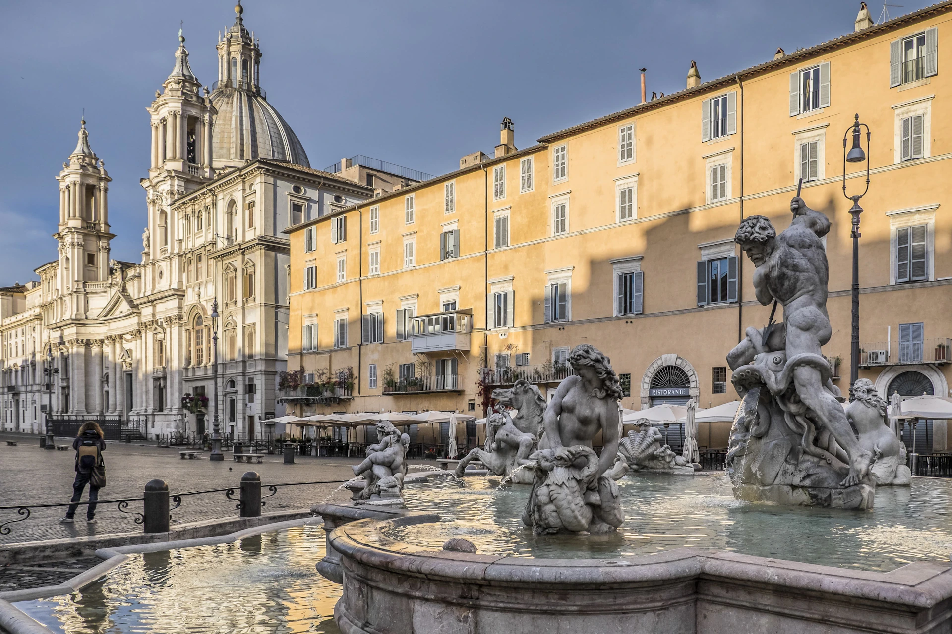 A historic European plaza with a Baroque church, yellow buildings, and a fountain featuring statues of human figures.