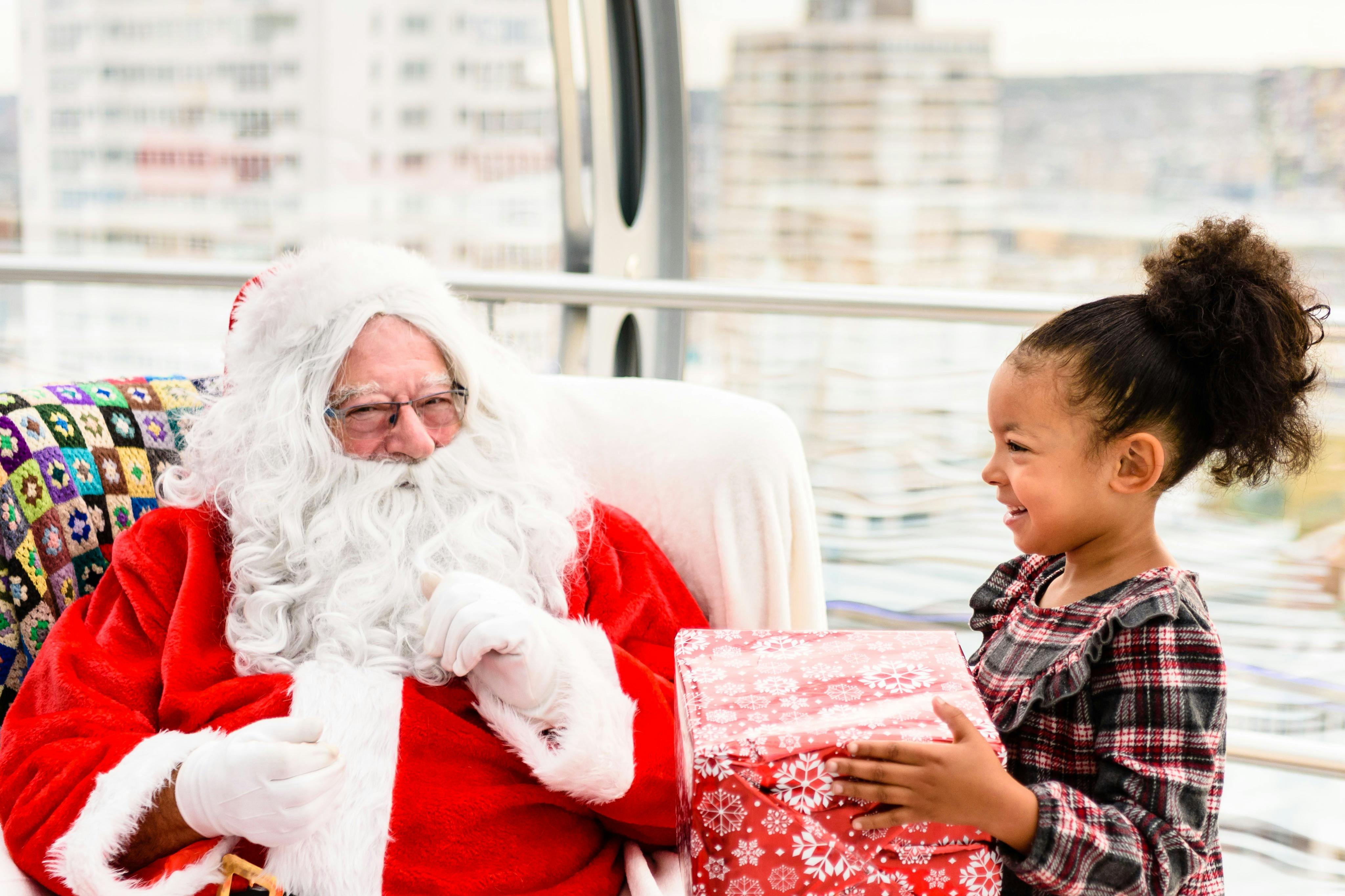 Santa Claus wearing his traditional red suit sits on a colorful blanket-covered chair, holding a red gift box.