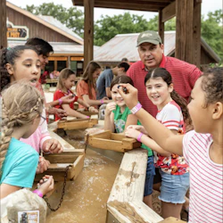Children and an adult engaged in gem mining activity at a wooden water trough, surrounded by rustic buildings and greenery.