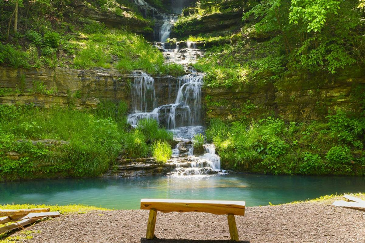 A wooden bench facing a cascading waterfall surrounded by lush greenery, with a turquoise pool at the base.