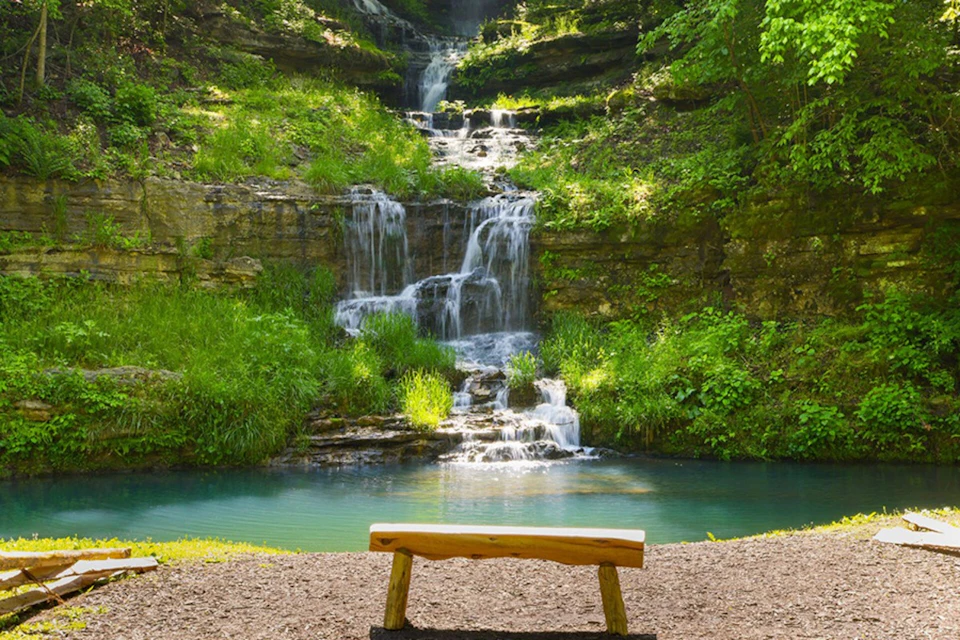 A wooden bench facing a cascading waterfall surrounded by lush greenery, with a turquoise pool at the base.