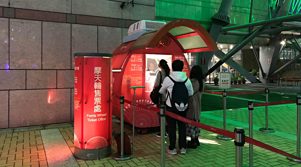 A red Ferris wheel ticket booth with customers standing in line, located indoors with signs and barriers around it.