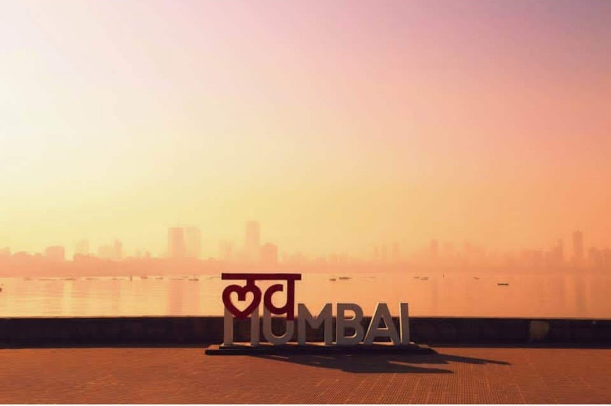 Panneau "I ❤️ Mumbai" au bord de l'eau au coucher du soleil, avec la ligne d'horizon de la ville en arrière-plan.