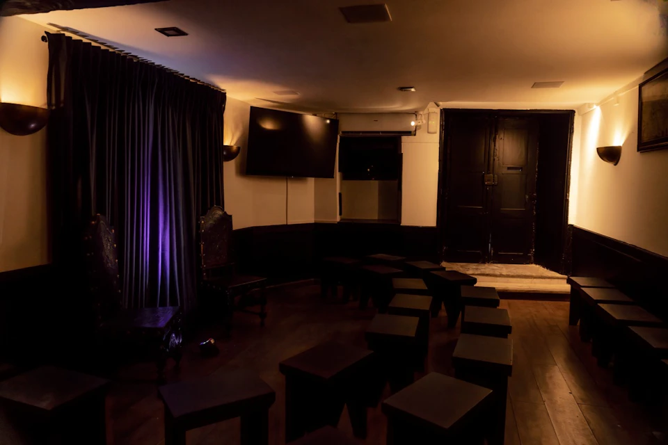 Dimly lit room with black curtains, small black tables arranged in rows, a wall-mounted TV, and a closed black door.