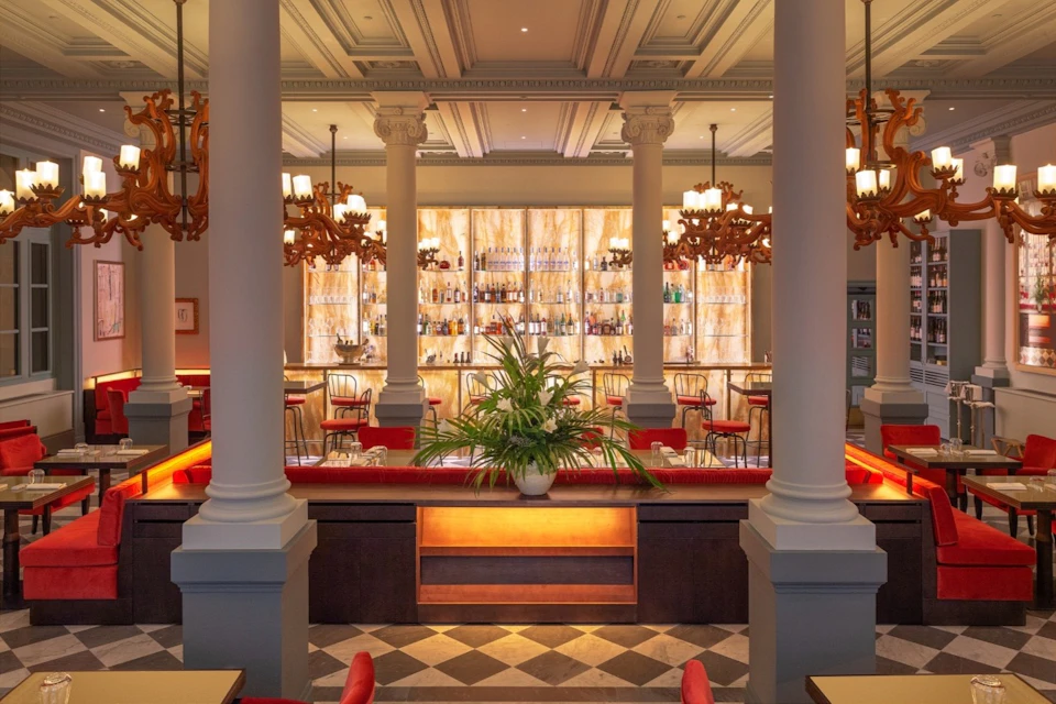Elegant bar with large columns, chandeliers, a backlit wall of bottles, red chairs, and a central plant in the foreground.