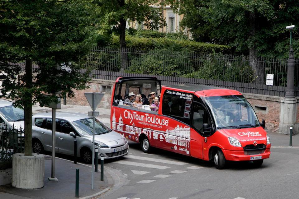 People are seated on a red open-top tour bus labeled "City Tour Toulouse" beside a silver car on a tree-lined street.