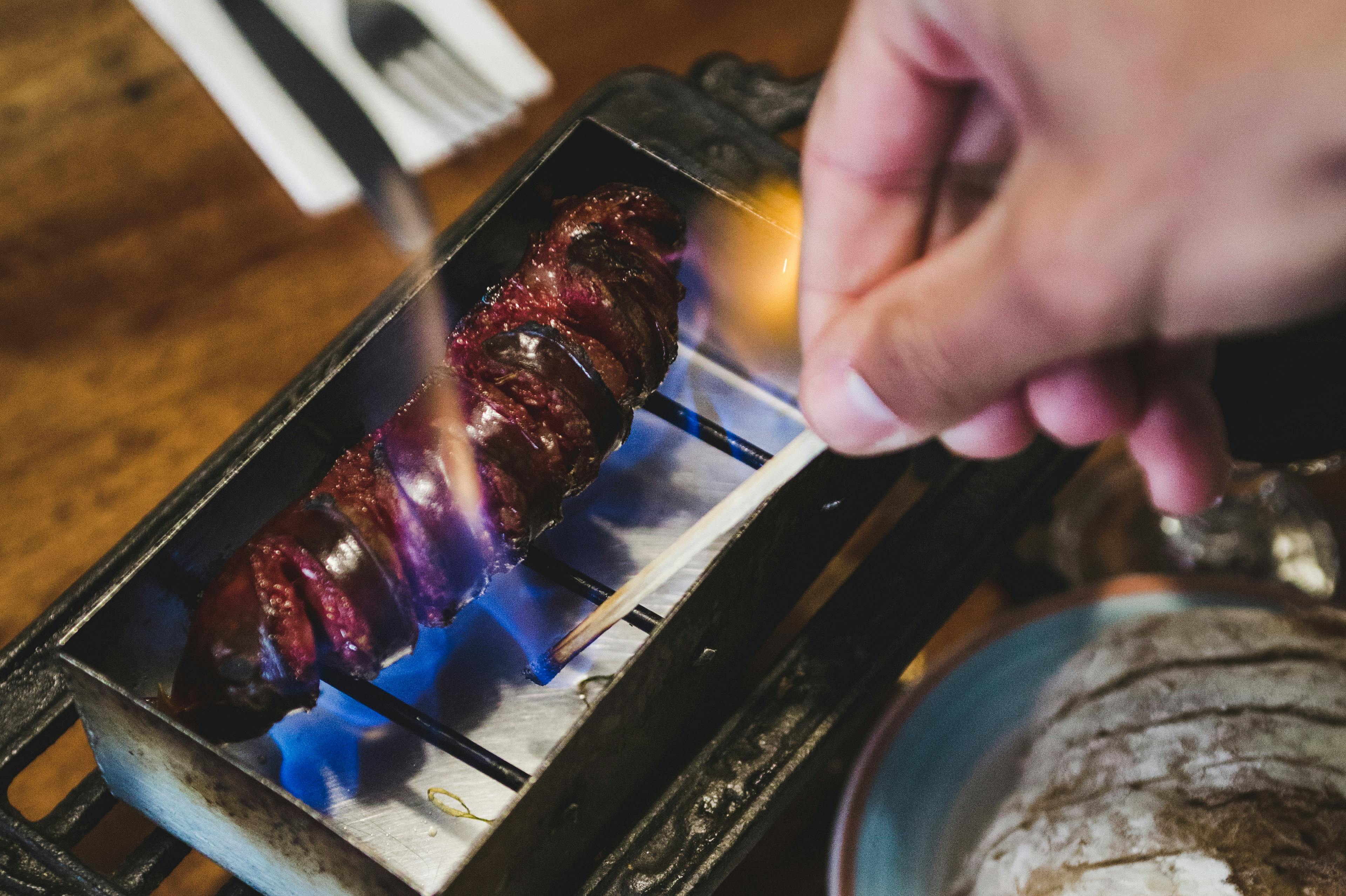 Iemand bakt een spies van spiraalvormig geroosterd vlees op een kleine tafelgrill met blauwe vlammen.