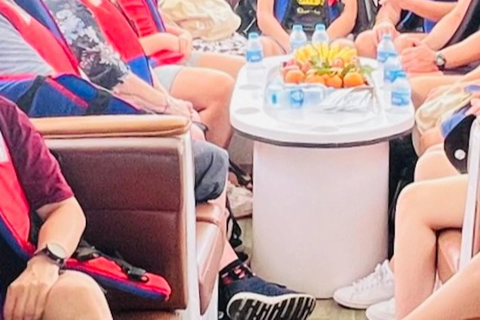 A group of smiling people wearing red life vests is seated inside a boat with a view of a cityscape visible through the window.