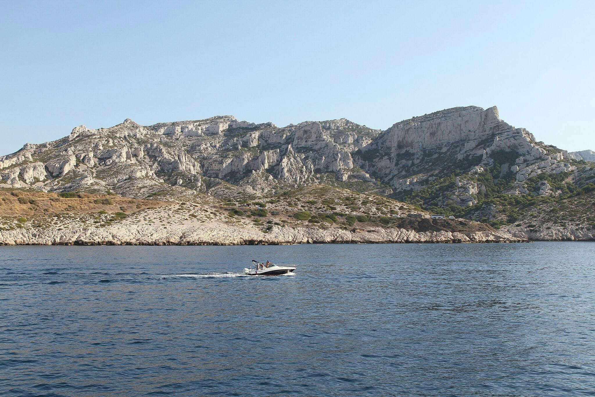 A motorboat with several people on board glides across a calm blue sea, with rocky hills and cliffs in the background under a clear sky.