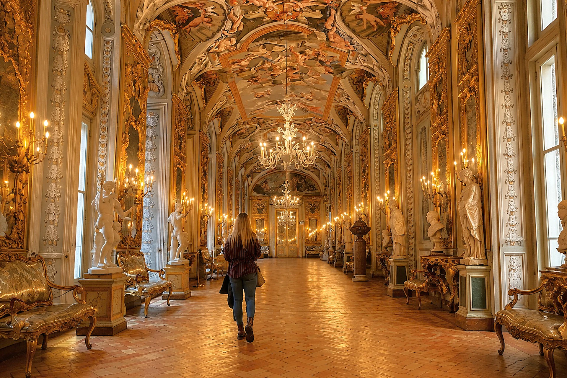 A woman walking through an ornate hall with chandeliers, statues, and intricate frescoes on the ceiling.