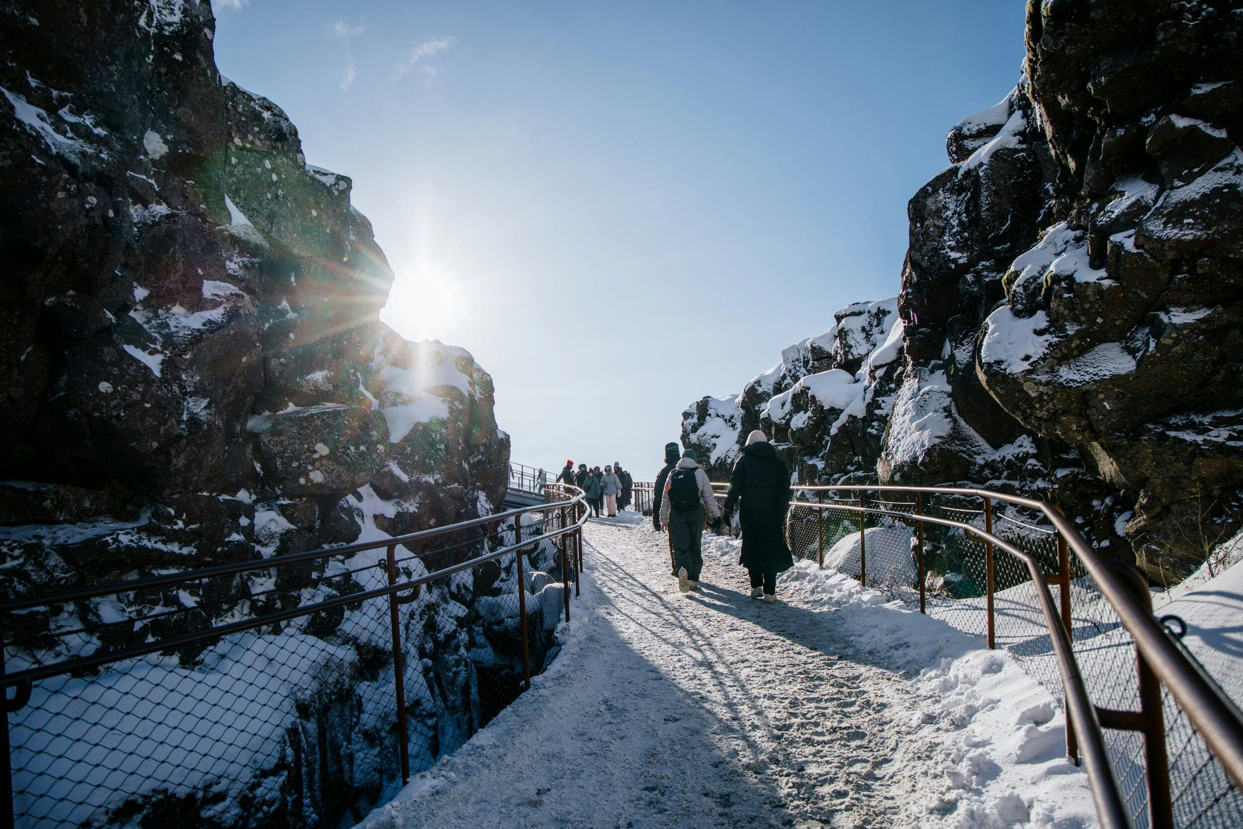 People walking on a snowy pathway between rocky cliffs under a bright sun and clear blue sky.