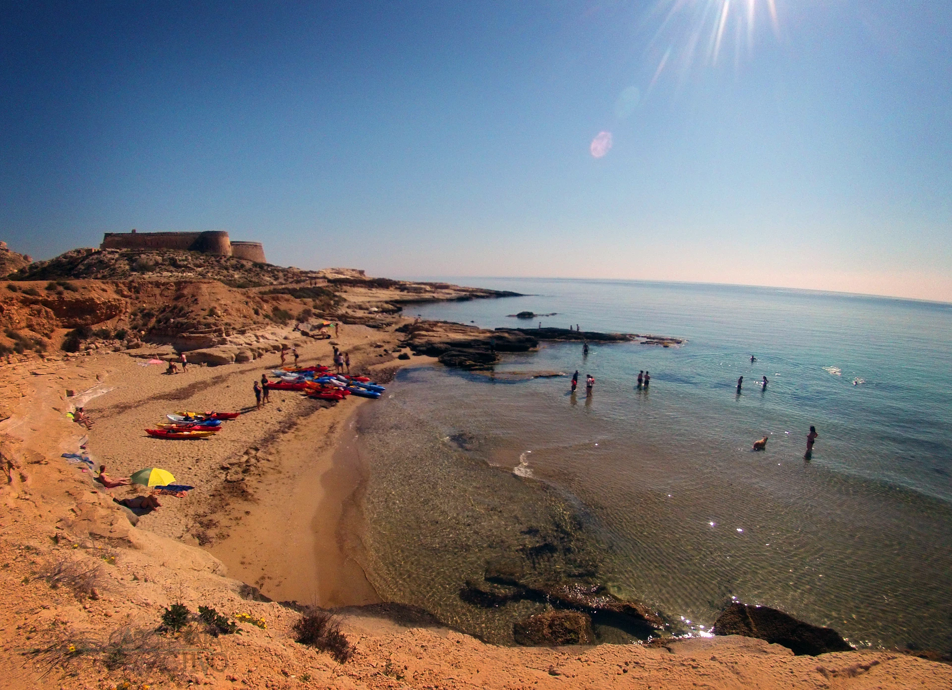 A sandy beach with kayaks and people scattered about in the shallow water. A rocky shore and a clear blue sky in the background.