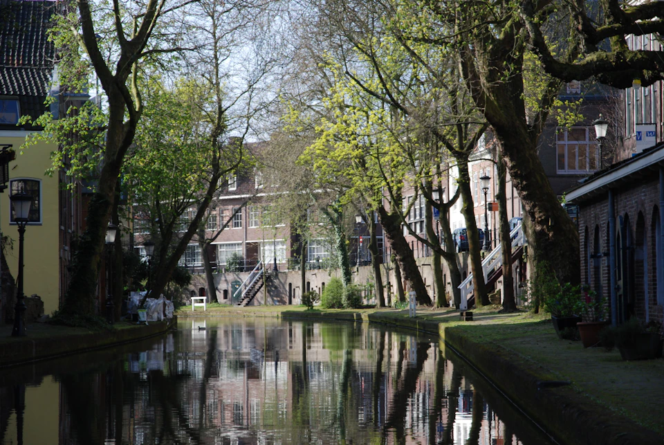 A canal lined with trees and historic buildings on a sunny day, with reflections in the calm water and stairs leading down to the riverbank.