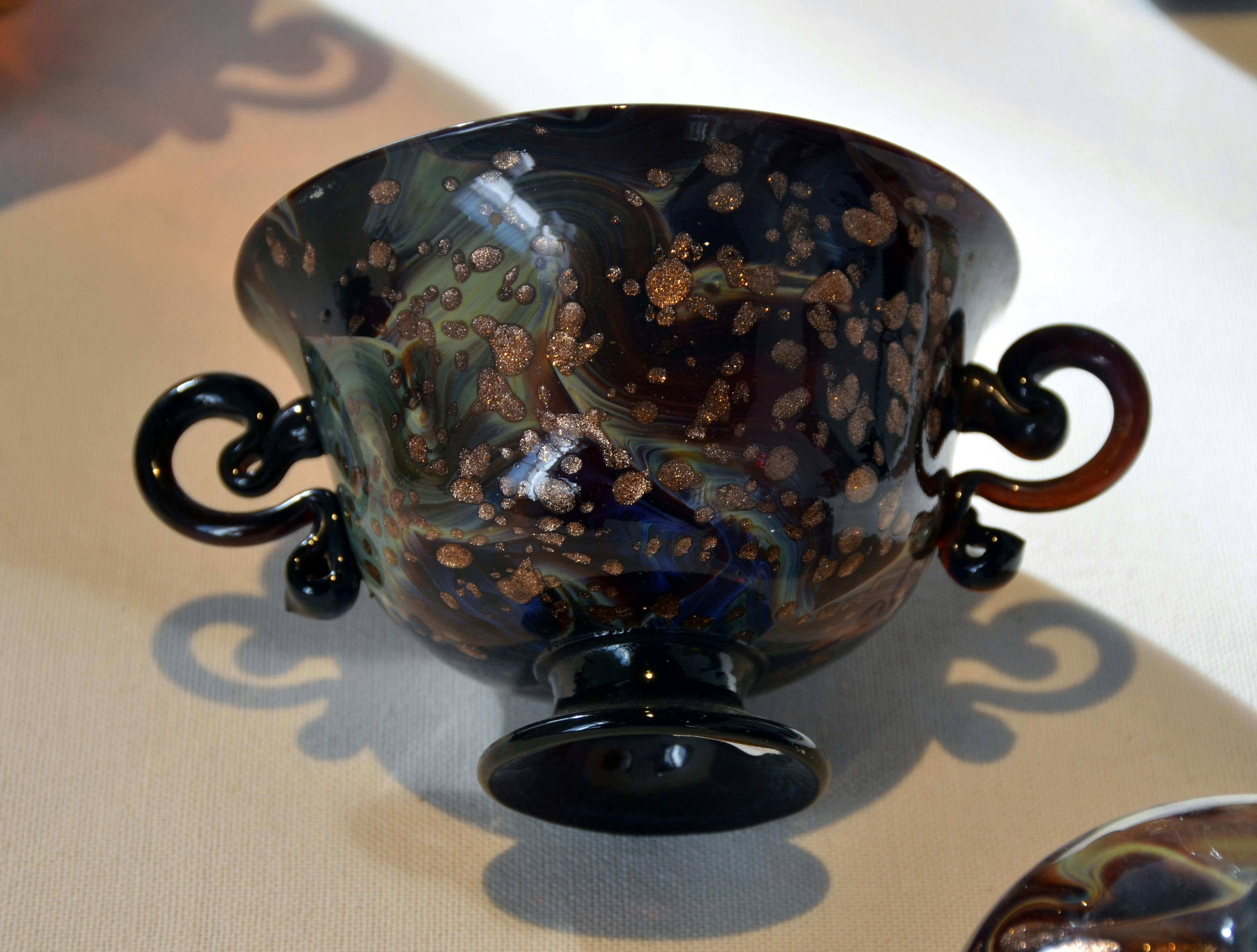 소용돌이치는 다양한 색상의 패턴과 양쪽에 두 개의 장식 손잡이가 있는 어둡고 화려한 세라믹 그릇입니다.