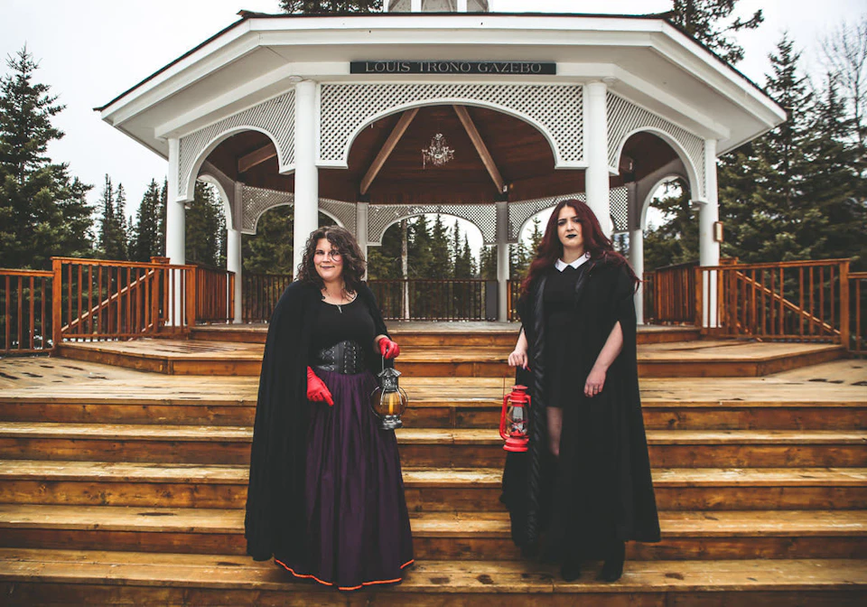 Two women in dark, vintage attire holding lanterns on wooden steps in front of a white gazebo labeled "Louis Trono Gazebo."