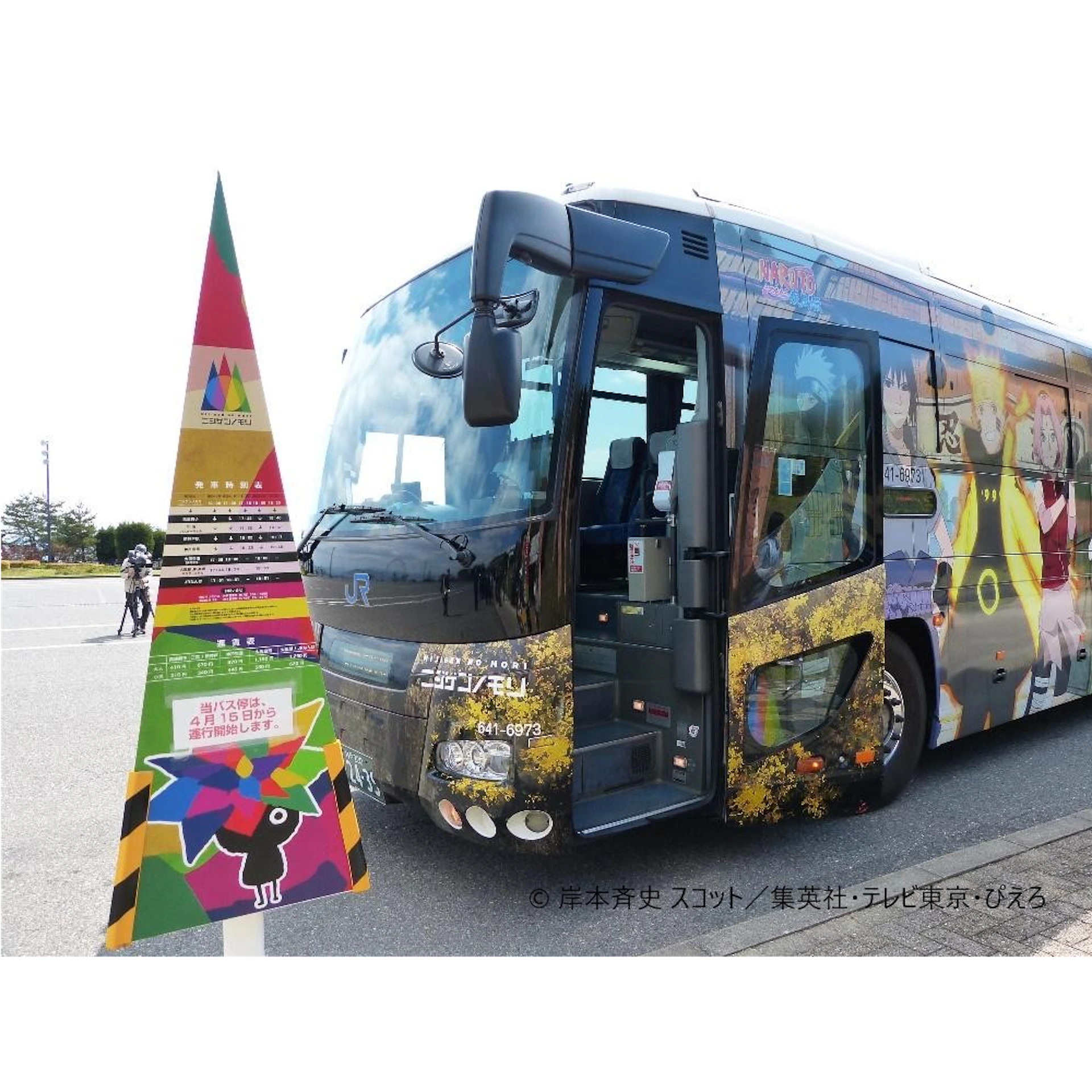 A colorful bus with animated character graphics is parked next to a tall, triangular sign with bright colors and text in Japanese.