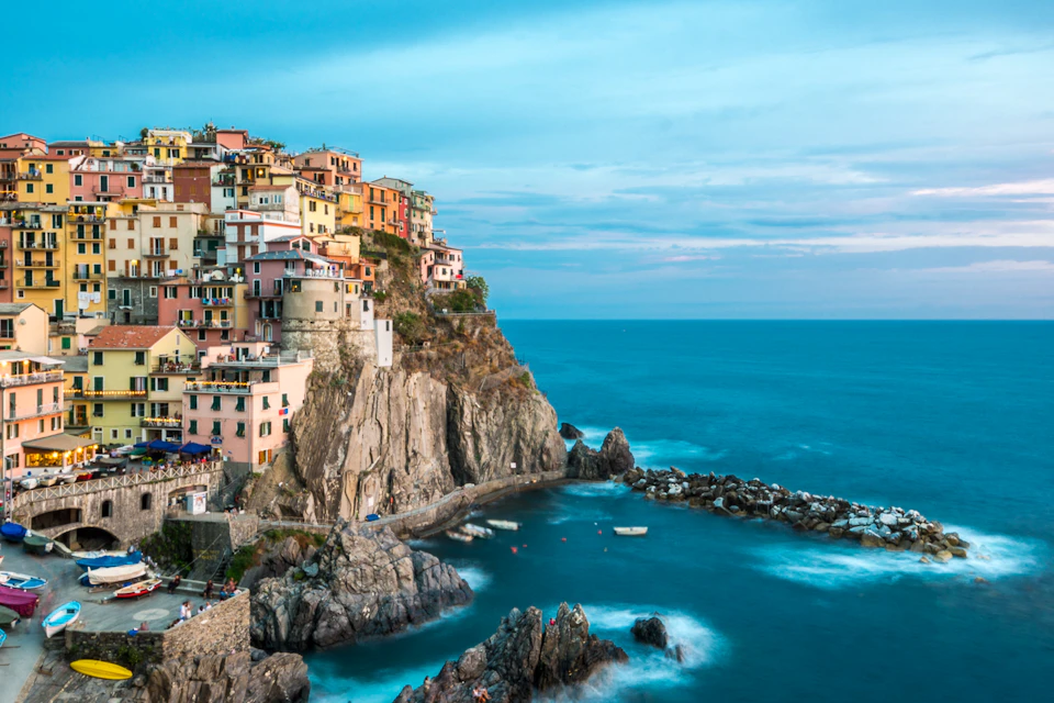 Colorful cliffside buildings overlooking a rugged coastline and blue sea under a partly cloudy sky. Small boats are in the water.