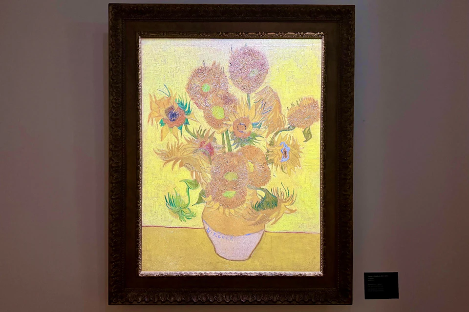 A painting of a vase with sunflowers, framed in an ornate wooden frame, displayed in a gallery setting.