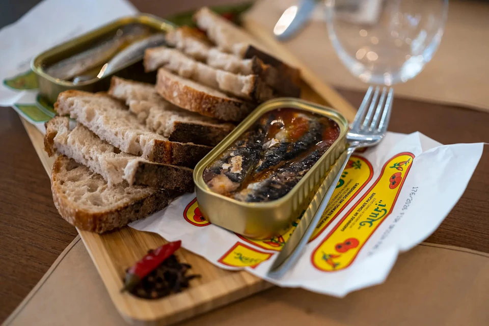 A wooden board with slices of bread and an open tin of sardines, accompanied by a fork and decorative packaging.