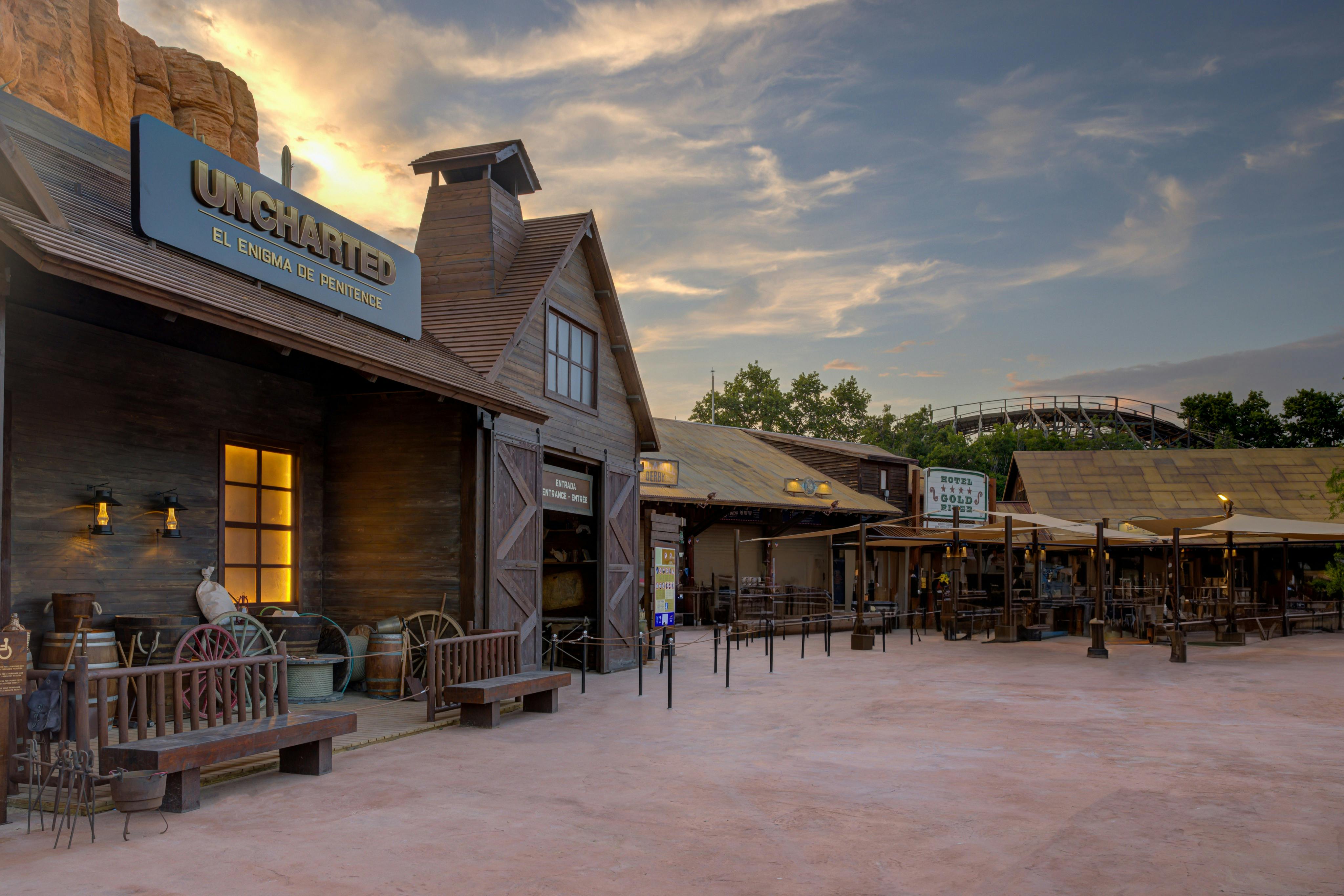 Des bâtiments à l'allure western, dont un magasin portant l'enseigne "UNCHARTED", sous un ciel parsemé de nuages au coucher du soleil.