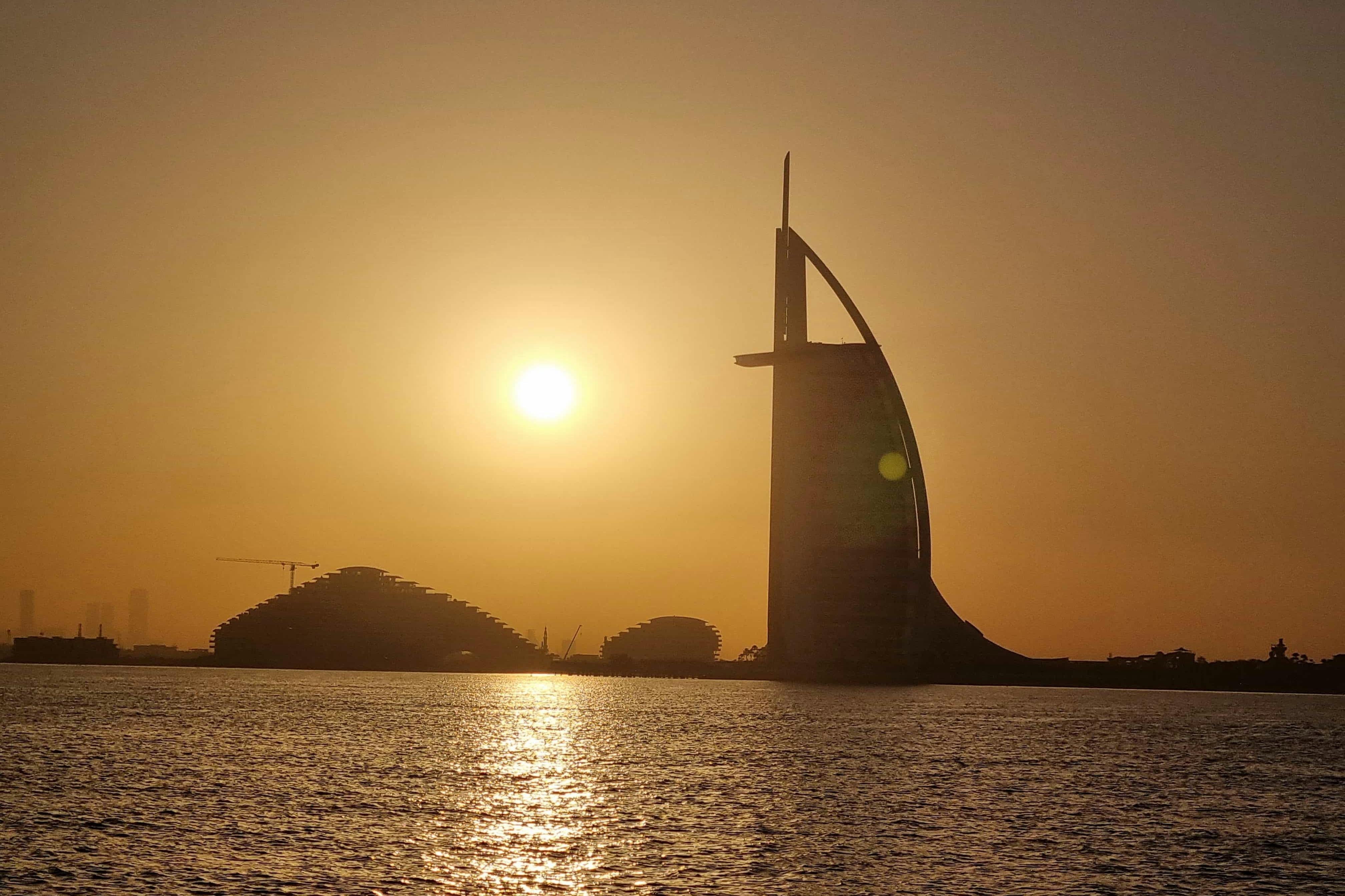 A silhouette of a sail-shaped building at sunset with calm water in the foreground.