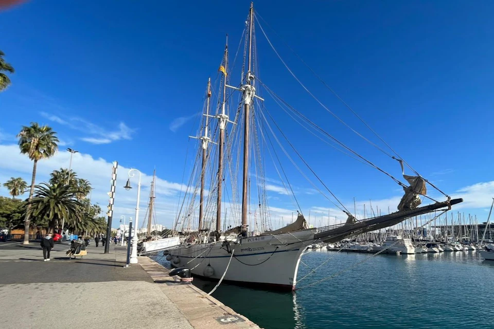 A large sailboat docked at a marina under a clear blue sky, with a walkway and lampposts along the pier.