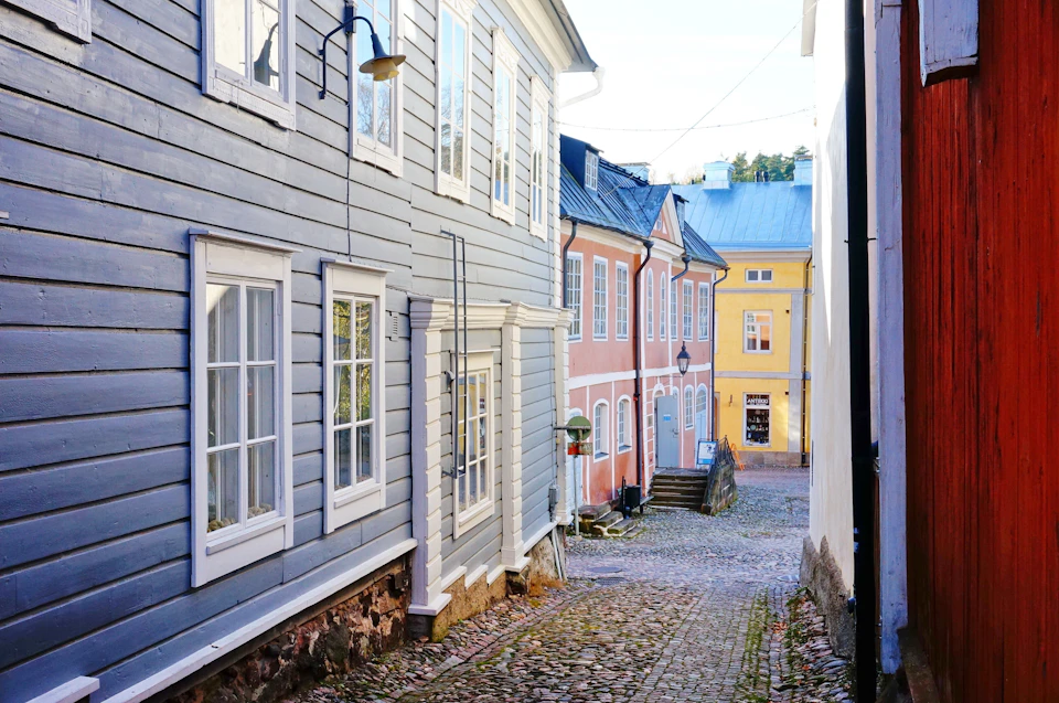 Narrow cobblestone street flanked by colorful wooden buildings with large windows and vintage lamps, leading to a yellow house.