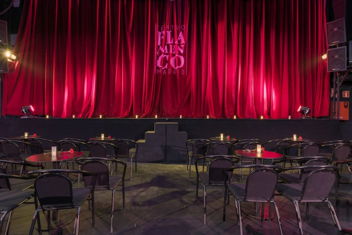Stage with red curtains in a dimly lit theater. Tables and chairs are arranged facing the stage, with candles on the tables.
