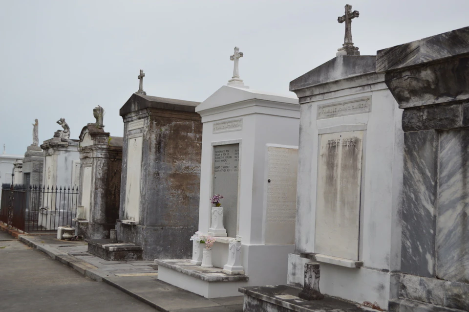 A row of tombs with crosses on top, some are weathered, one is white and decorated with flowers, under a cloudy sky.