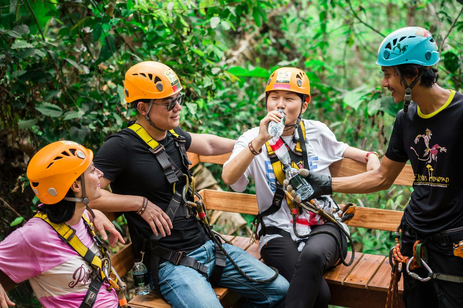 三个戴着头盔和马具的人坐在郁郁葱葱的森林里的木凳上，喝着水，面带微笑。