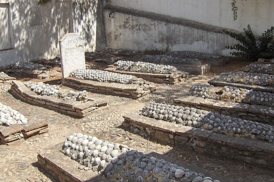 Several graves with tombstones covered by shells in an old cemetery with a white wall in the background.