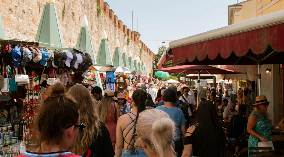 Crowded outdoor market with people browsing stalls against an old brick wall, selling hats, clothes, and accessories.