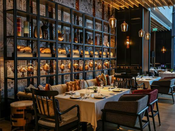 A cozy restaurant interior with tables set for dining, decorative shelves, and warm ambient lighting.
