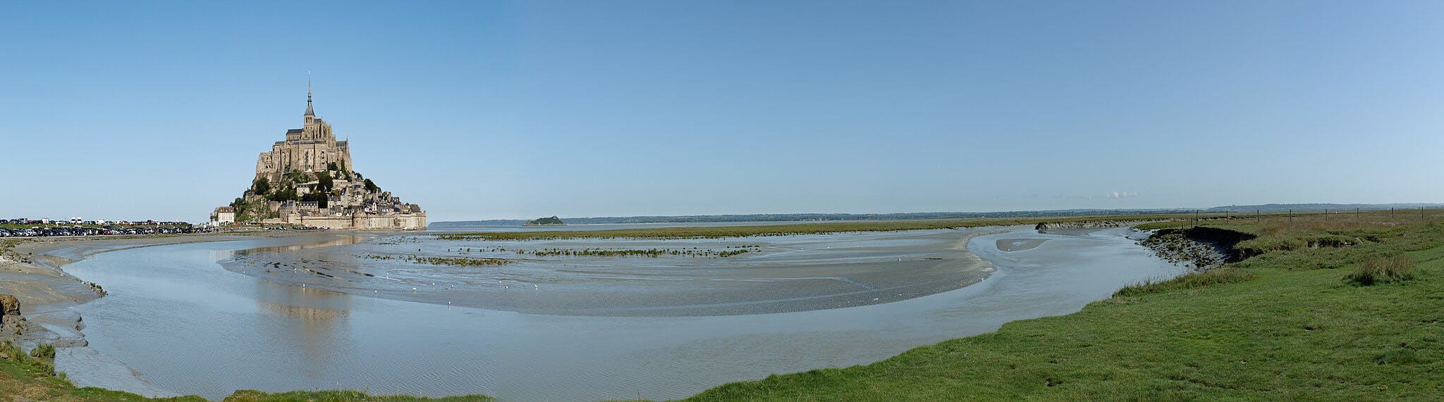 Uma abadia medieval numa ilha rochosa rodeada por águas de maré, com um primeiro plano relvado e um céu azul claro.