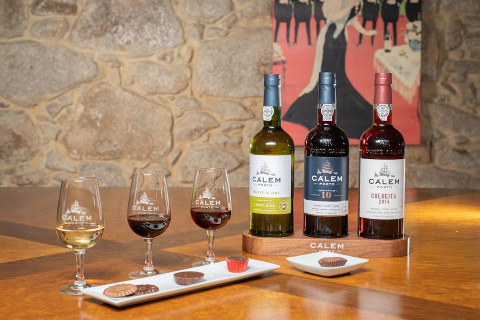 Three bottles of Calem Porto wine, glasses filled with wine, and plates with chocolates arranged on a wooden table.