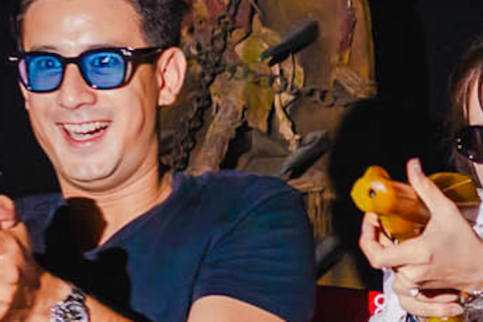 Two people seated in a red amusement ride car, holding toy guns, surrounded by skeleton props in a dark setting.