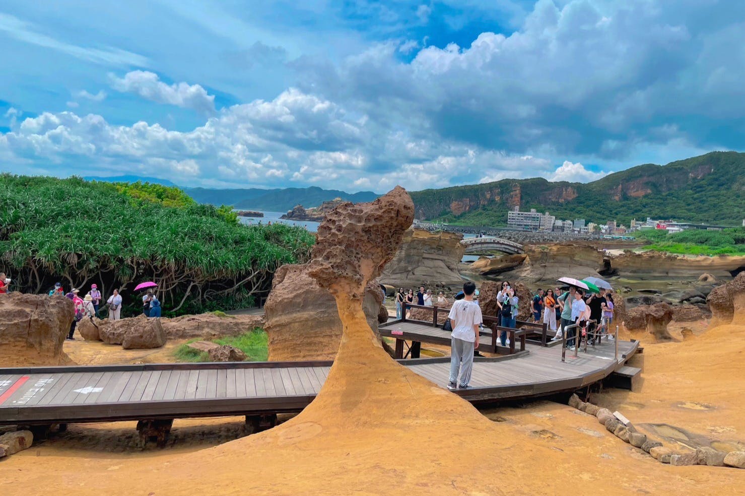 Tourists with umbrellas walk on a wooden path near a unique rock formation with green hills and cloudy skies in the background.