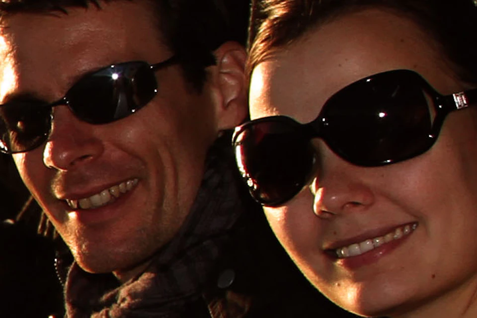 Two people in sunglasses smile on a boat at sunset with a large metal bridge in the background.