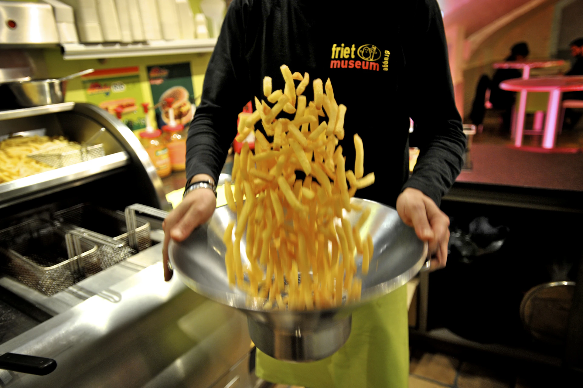 Person tossing a bowl of French fries at the Friet Museum. Background includes kitchen equipment and colorful posters.