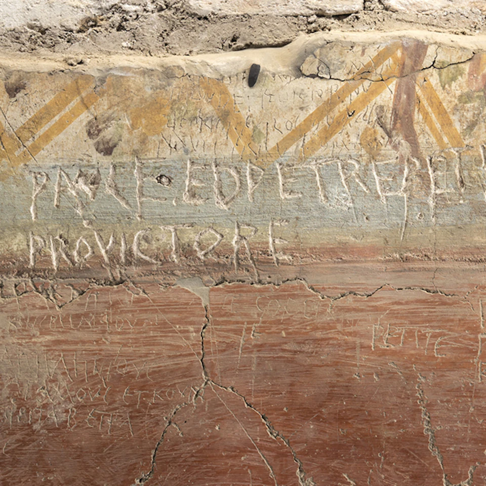 Ancient wall with faded fresco and Latin graffiti, showing geometric patterns and various etched inscriptions. Cracks visible in surface.