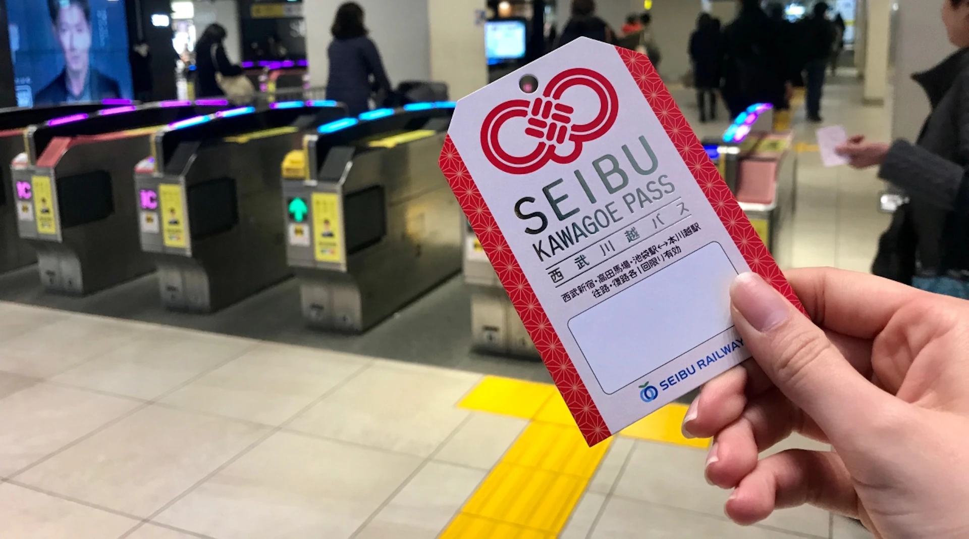 A hand holding a Seibu Kawagoe Pass ticket in front of a train station ticket gate area.
