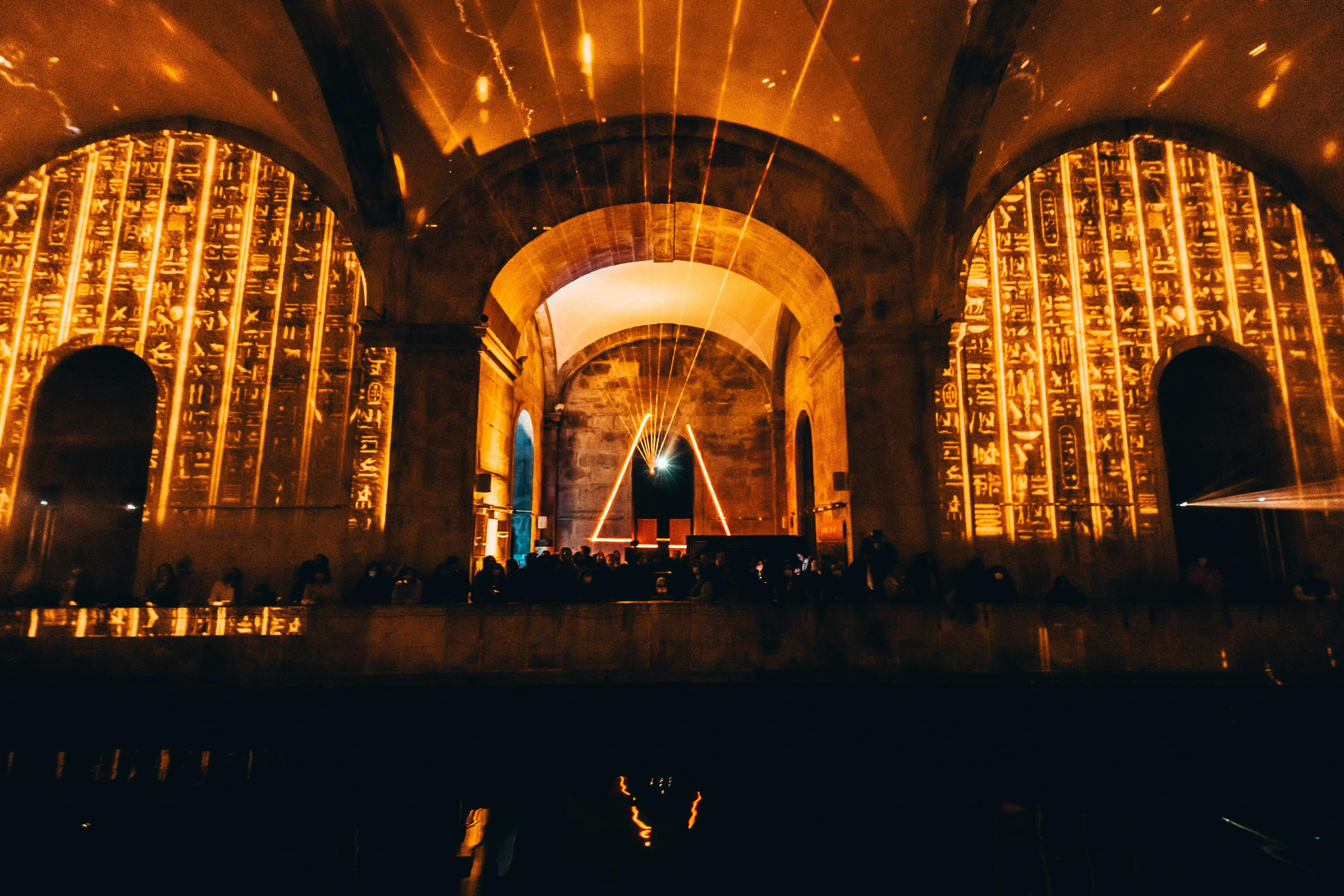 石のアーチ、照らされた象形文字のパネル、中央に三角形のライトディスプレイがある薄暗い部屋。シルエットの人。