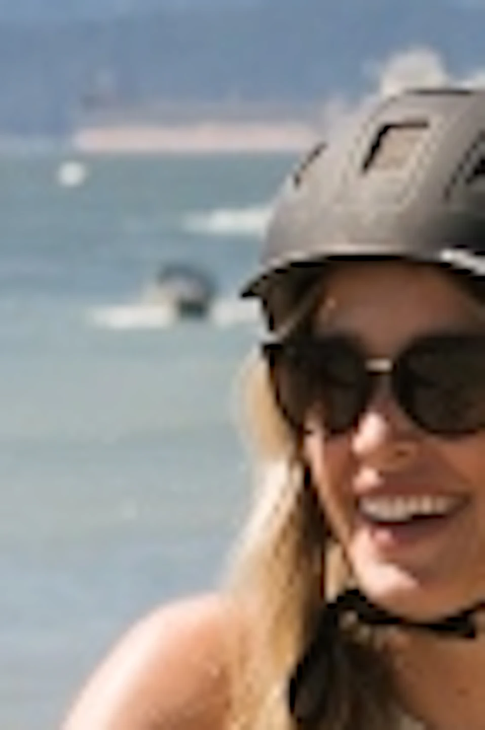 Three people wearing helmets walk their bicycles on a sandy beach with mountains and boats visible in the background.