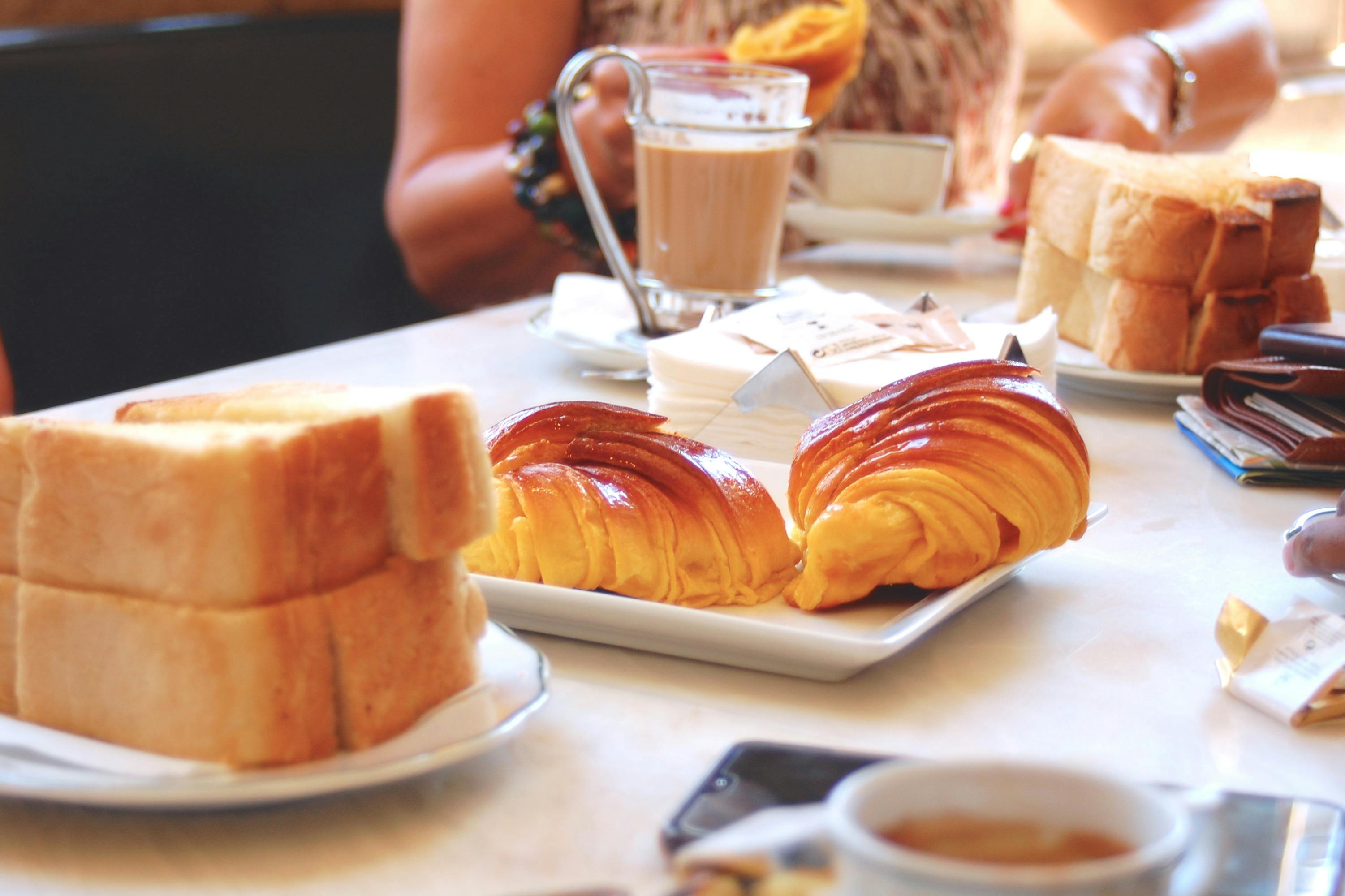Ένα τραπέζι για πρωινό με κρουασάν, ψωμί σε φέτες, καφέ και ανθρώπους στο παρασκήνιο που κρατούν γλυκά και ποτά.