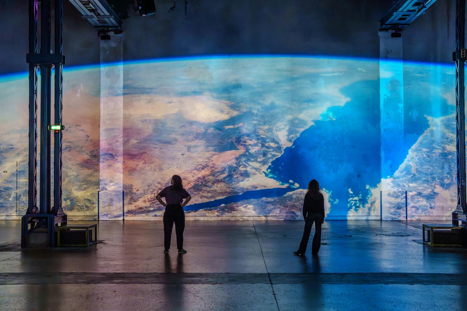 Two people stand facing a large, wall-sized image of Earth as viewed from space, featuring continents and oceans in vivid colors.